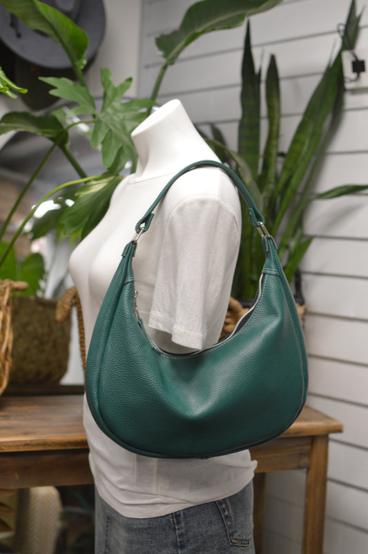 A teal green pebbled leather hobo shoulder bag with silver-tone hardware, displayed on a white mannequin torso in a styled indoor setting with plants and a wooden table.